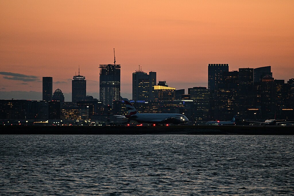 British Airways plane landing in Boston with water in front and a cityscape and sunset behind.