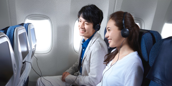 Two passengers are enjoying in-flight entertainment with headphones on, smiling and relaxed, during an ANA flight.