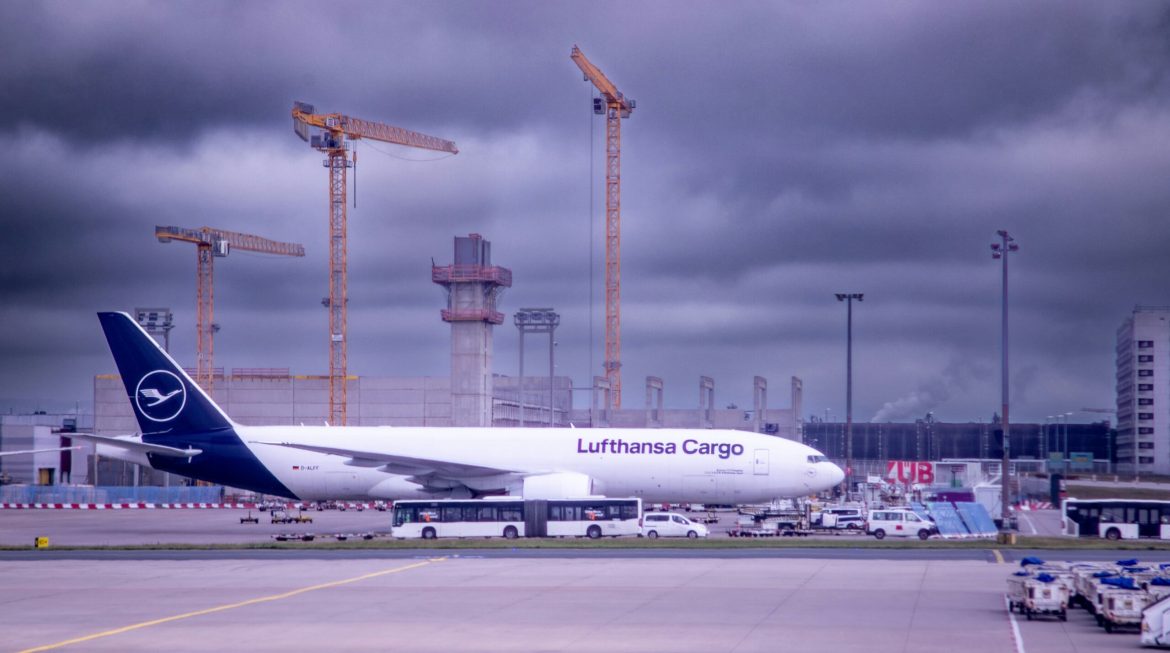 A Lufthansa Cargo Boeing 777F cargo plane is parked on the tarmac at an airport, with construction cranes and terminal buildings in the background. Ground support vehicles and crew are visible around the aircraft, highlighting the busy operations of air freight logistics. The overcast sky adds a dramatic backdrop to the scene, emphasizing the scale and importance of cargo transportation in the aviation industry.