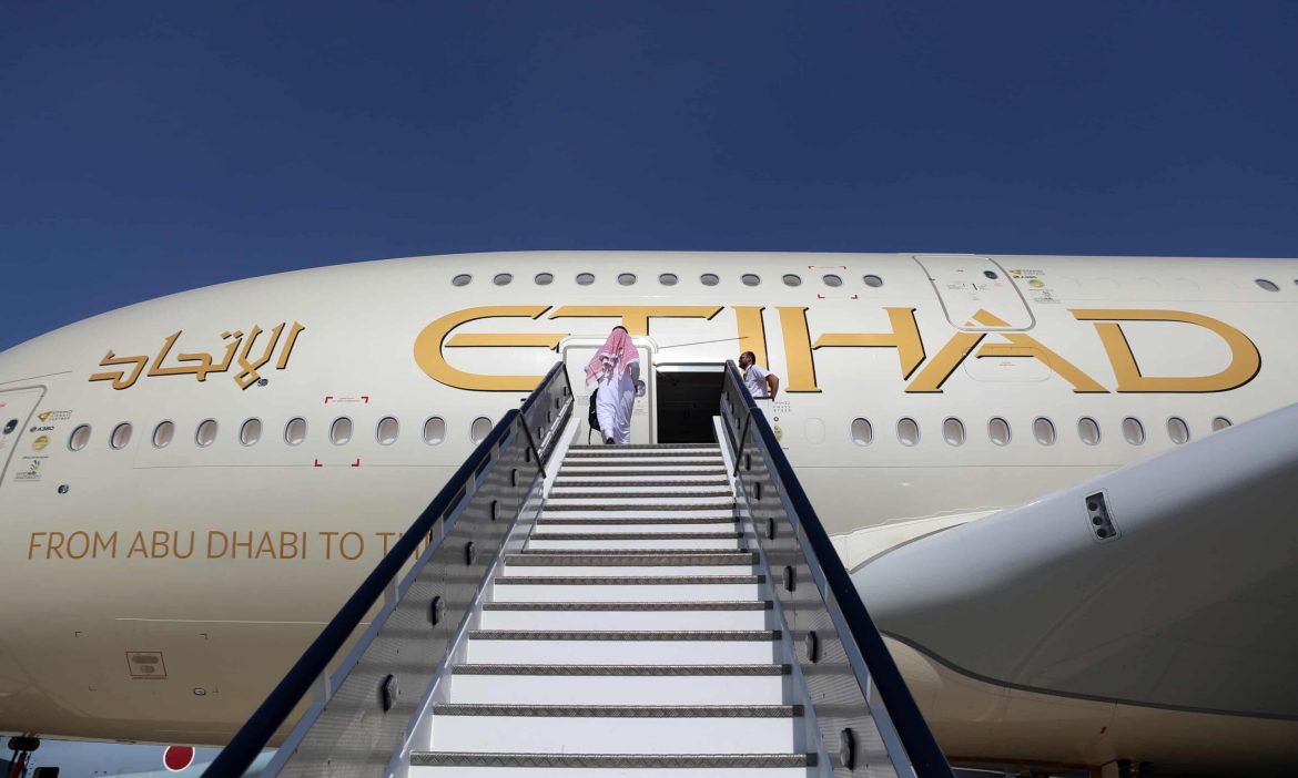 An Etihad plane is parked and has its door opened with stairs up to it. An indivual is at the top of the stairs.