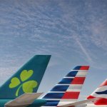 The tails of an Aer Lingus, American Airlines and British Airways planes sit on a runway in front of a blue sky.