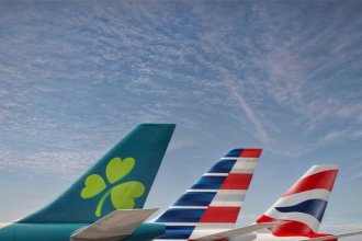 The tails of an Aer Lingus, American Airlines and British Airways planes sit on a runway in front of a blue sky.