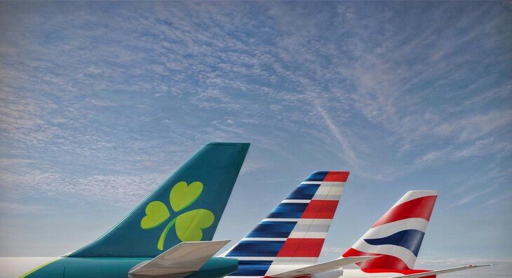 The tails of an Aer Lingus, American Airlines and British Airways planes sit on a runway in front of a blue sky.