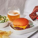 A photoshoot of the inflight meal that includes a cheeseburger, Caesar salad, fires and brownie. Also included in the photo is a glass of beer and a hand holding the brownie.