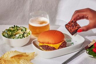 A photoshoot of the inflight meal that includes a cheeseburger, Caesar salad, fires and brownie. Also included in the photo is a glass of beer and a hand holding the brownie.