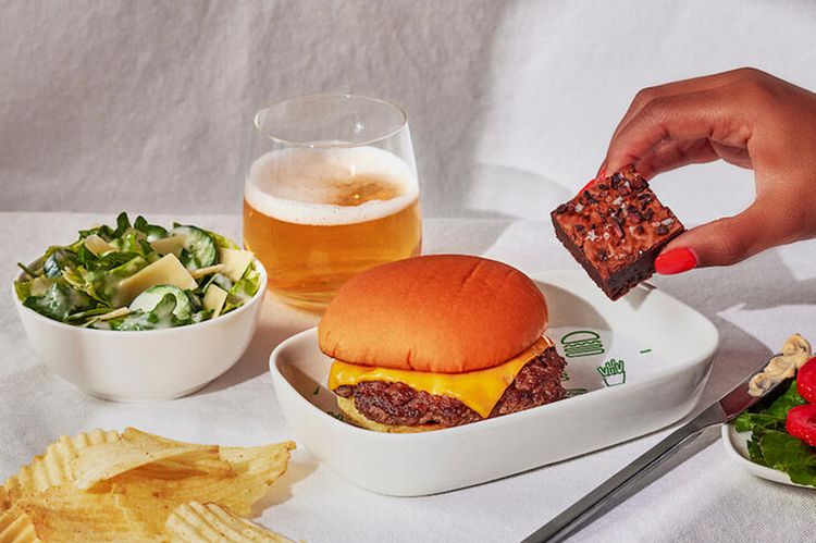 A photoshoot of the inflight meal that includes a cheeseburger, Caesar salad, fires and brownie. Also included in the photo is a glass of beer and a hand holding the brownie.